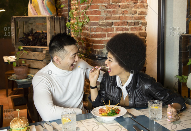 Laughing African American young woman feeding cheerful Asian boyfriend with salad while having fun during lunch in cafe