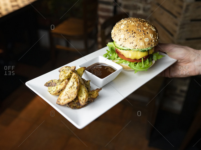 Crop anonymous waiter holding tray with delicious appetizing burger served with baked potatoes and sauce while serving meal for clients in cafe