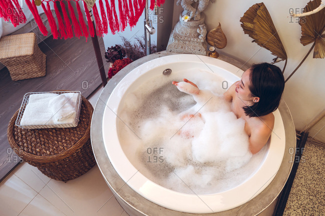 From above of relaxed young Asian female sitting in small round bathtub with foam and enjoying bath procedure in spa salon with oriental decor