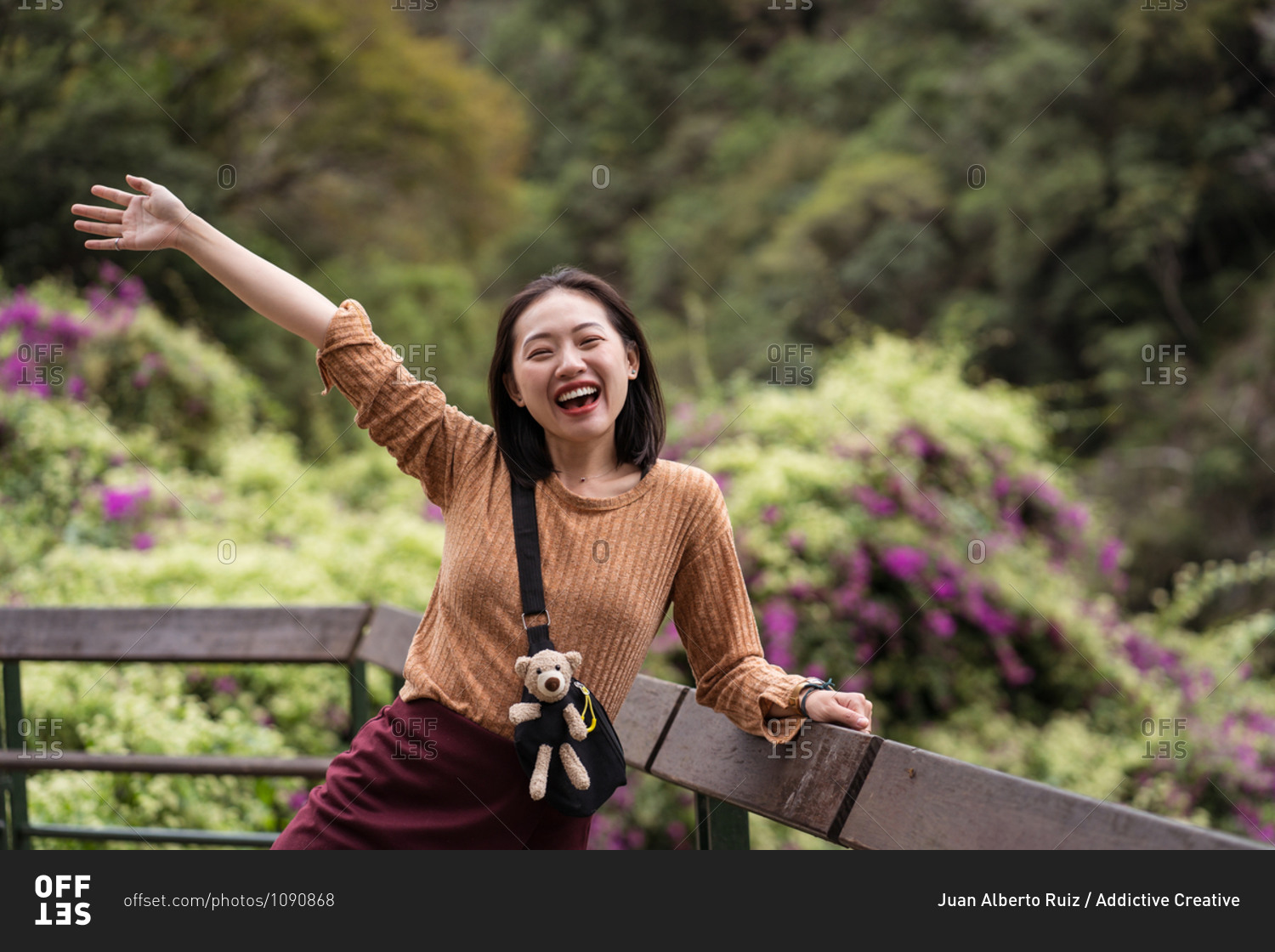 Cheerful Asian female tourist standing on wooden terrace with raised arm while enjoying summer vacation in green park in Hualien City and looking at camera
