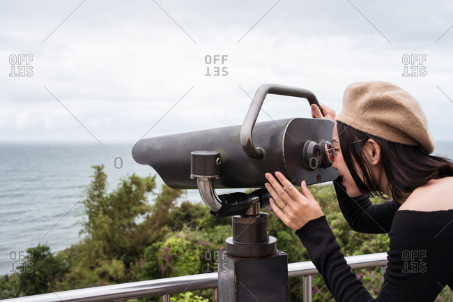 Side view of cheerful young Asian female tourist enjoying view of sea through binocular while standing on observation point during trip to Taiwan