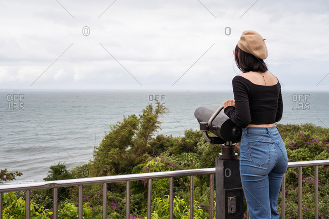 young Asian female traveler looking at camera while standing near binocular on observation point against sea and cloudy sky in Taiwan