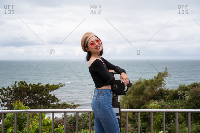 Side view of smiling young Asian female traveler looking at camera while standing near binocular on observation point against sea and cloudy sky in Taiwan