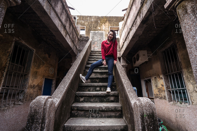Low angle full body of positive young Asian female traveler in casual clothes standing on stone stairway of aged abandoned house while exploring streets of Kaohsiung city in Taiwan