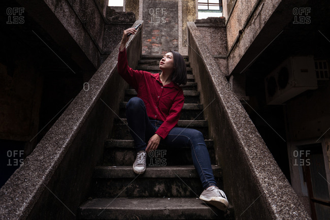 Low angle full body of young Asian female traveler in casual clothes sitting on stone stairway of aged abandoned house and taking selfie while exploring streets of Kaohsiung city in Taiwan