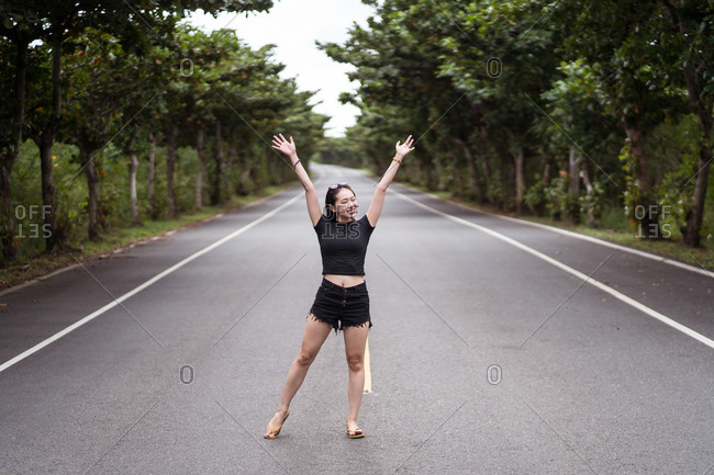 Full body of positive young Asian female traveler in casual wear looking away while standing on empty asphalt path among green trees in Kenting National park in Taiwan