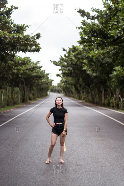 Full body of positive young Asian female traveler in casual wear looking at camera while standing on empty asphalt path among green trees in Kenting National park in Taiwan