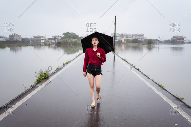 Melancholic female with umbrella walking barefoot along wet empty road on rainy day under cloudy sky in Yilan County