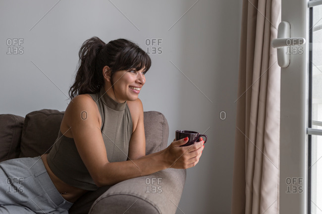 Side view smiling content female in casual wear relaxing on comfy sofa with cup of hot drink and looking out window