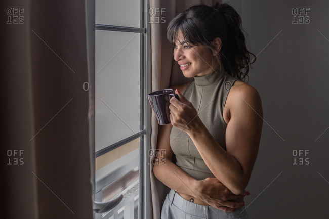 Side view smiling content female in casual wear relaxing standing by a window with cup of hot drink and looking out