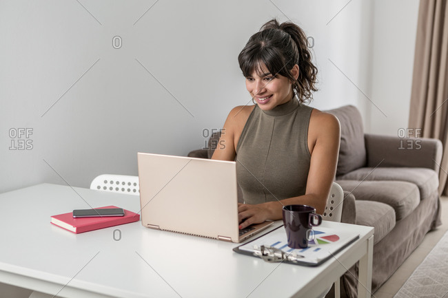 Optimistic smiling female sitting at desk working on netbook in living room at home in modern apartment