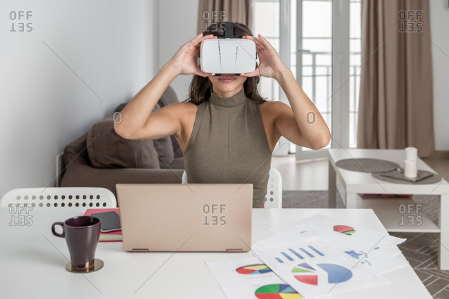 Positive smiling female with arms raised using VR headset while sitting at table with contemporary laptop and papers