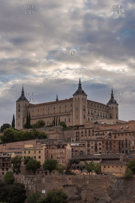 Aerial view of old city with historic buildings and stone castle on hill against cloudy sky during sunset in Toledo, Spain