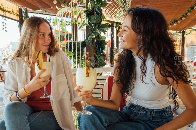 Happy young female friends sitting in cozy outdoor cafe and toasting with glasses of fresh fruit drinks while spending summer day together