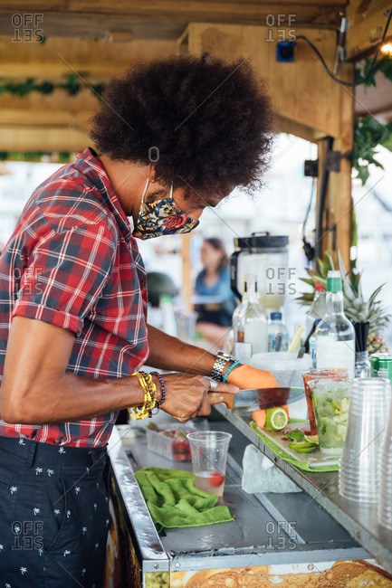 African American male barkeeper in protective mask garnishing cocktail with slice of fresh lime while preparing drink at counter of beach bar