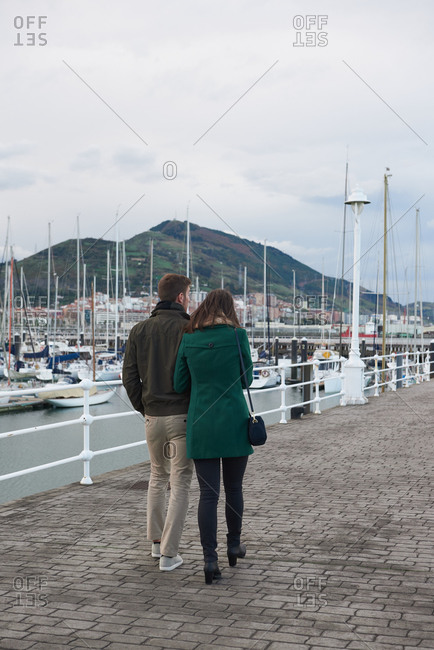 Back view of couple holding hands and walking along embankment in port near sea while enjoying stroll in autumn together