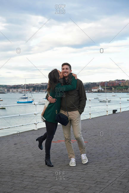 Delighted woman kissing smiling man in cheek while standing at seafront in autumn and enjoying stroll
