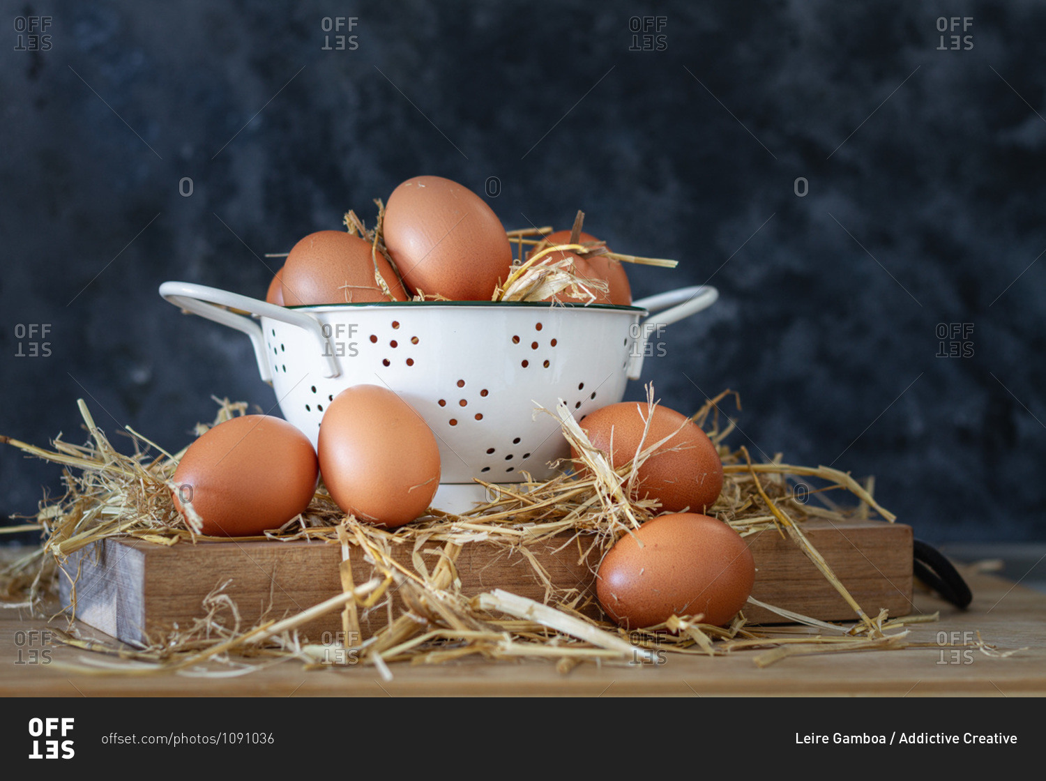 Composition with brown chicken eggs in white bowl placed on wooden board with dry hay against dark background