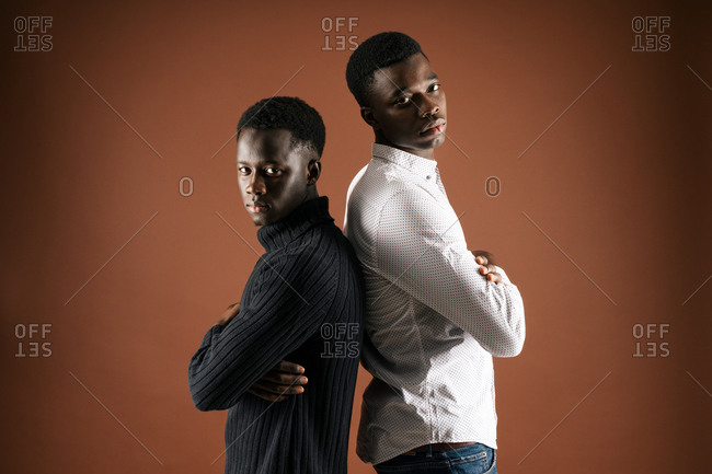 Side view of determined African American male friends standing back to back with crossed arms on brown background and looking at camera