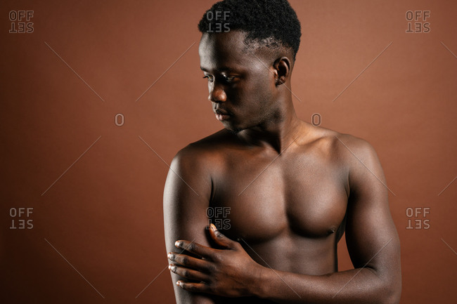 Confident African American young male with muscular naked torso standing in trendy jeans with hand on waist on brown background in studio and looking at camera