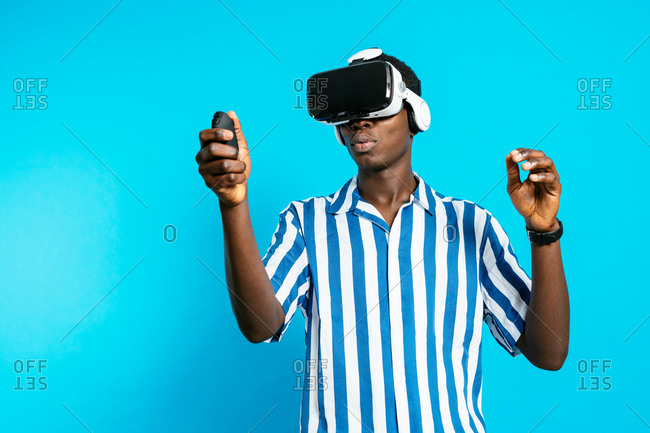 Side view of young African American male in VR goggles and with joystick experiencing virtual reality on blue background in modern studio
