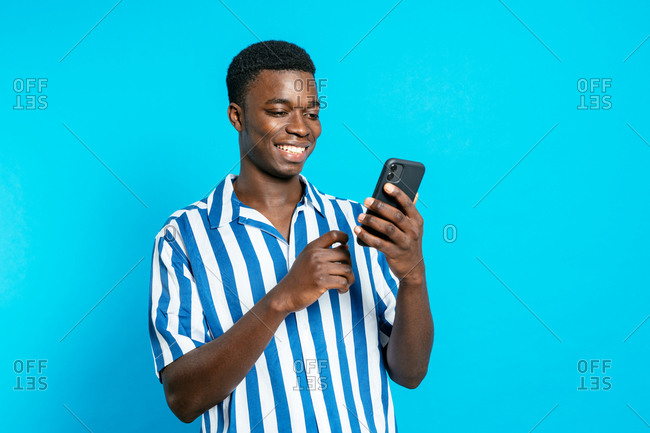 Delighted African American male browsing smartphone and reading good news about business project while standing with clenched fist on vivid blue background in studio