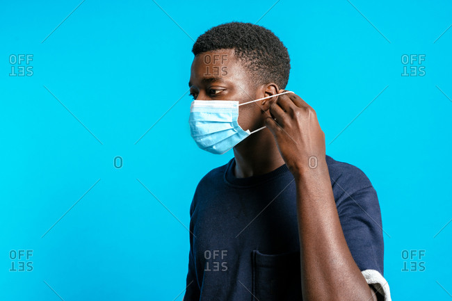 Serious African American male putting on sterile medical mask while standing on bright blue background in studio and showing concept of coronavirus prevention