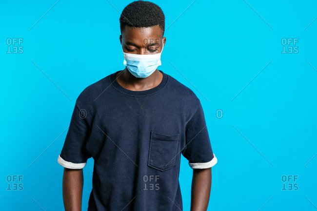 Serious African American male with sterile medical mask while standing on bright blue background in studio and showing concept of coronavirus prevention