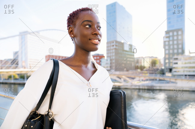 Confident successful positive young black businesswoman with short hairstyle standing near river against modern buildings in downtown