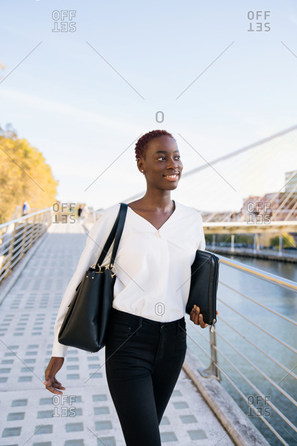 Confident successful positive young black businesswoman with short hairstyle standing near river against modern buildings in downtown