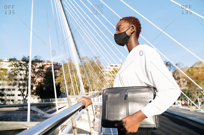 Low angle of stylish confident young African American businesswoman in black protective mask with handbag and laptop case standing on urban street with contemporary buildings