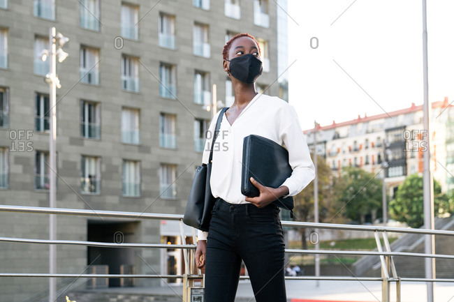 Low angle of stylish confident young African American businesswoman in black protective mask with handbag and laptop case standing on urban street with contemporary buildings