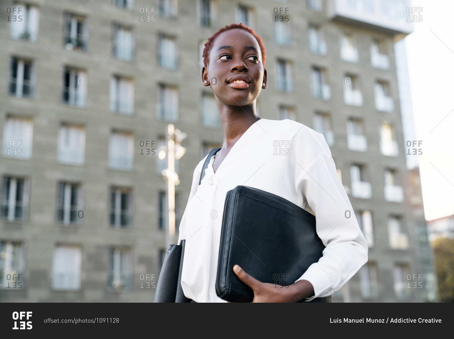 Low angle side view of confident smiling young African American female manager in elegant white blouse carrying laptop case and looking away while standing near modern urban building