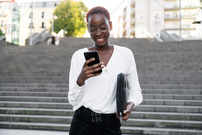 Young African American businesswoman in classy outfit reading message on smartphone and smiling satisfied with news while standing on stairway on urban street