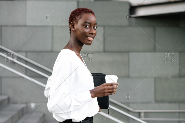 Side view of positive elegant young black business lady with cup of takeaway coffee standing near modern urban building