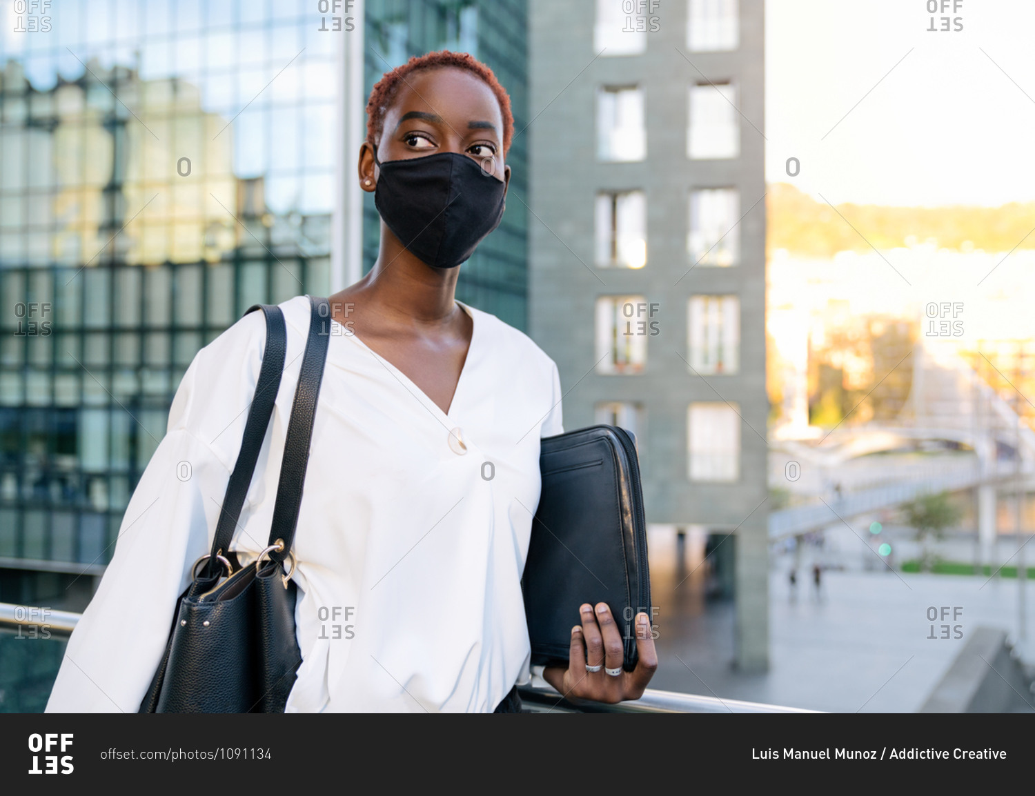 Serious stylish young African American business lady in white blouse and black protective mask holding case with laptop while standing against contemporary glass building in modern city district