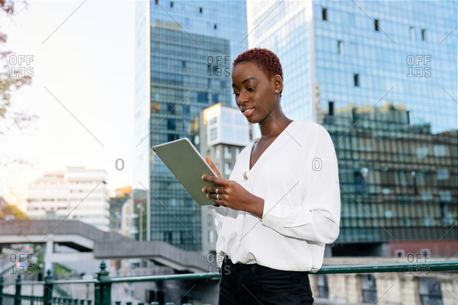Contemporary African American female entrepreneur in stylish outfit browsing tablet while standing against modern glass building in city