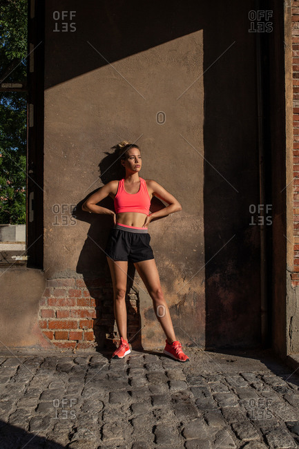 Fit female athlete in activewear leaning on wall of shabby building and looking away while standing in city during training