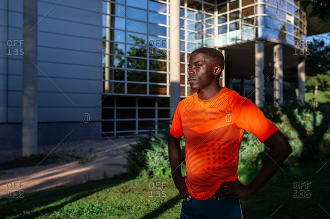 Determined African American male athlete in sportswear standing with hands on waist in urban area of city and looking away