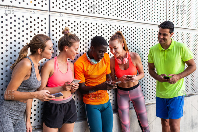 Group of multiracial runners in activewear leaning on wall of building and using smartphones after workout in city