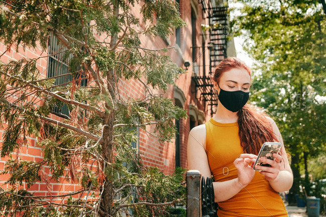 Young woman on phone wearing facemask in brooklyn street by trees.