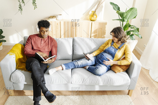 Husband listening to music and reading a book with his pregnant wife who is watching a tablet on a sofa. interracial couple concept