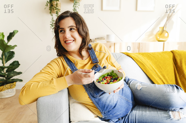 Pregnant young woman smiling and eating a salad on a sofa. maternity concept