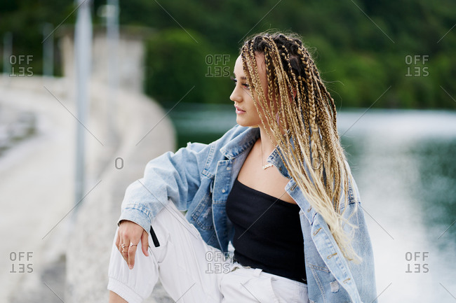 Young woman with blonde braided hair wearing a denim jacket and white jean resting on a bridge