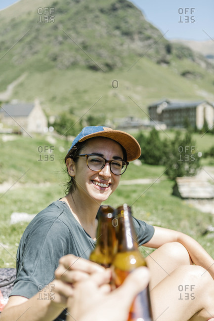 Traveler woman toasting with beer with anonymous man while sitting by the side of a lake in the mountains