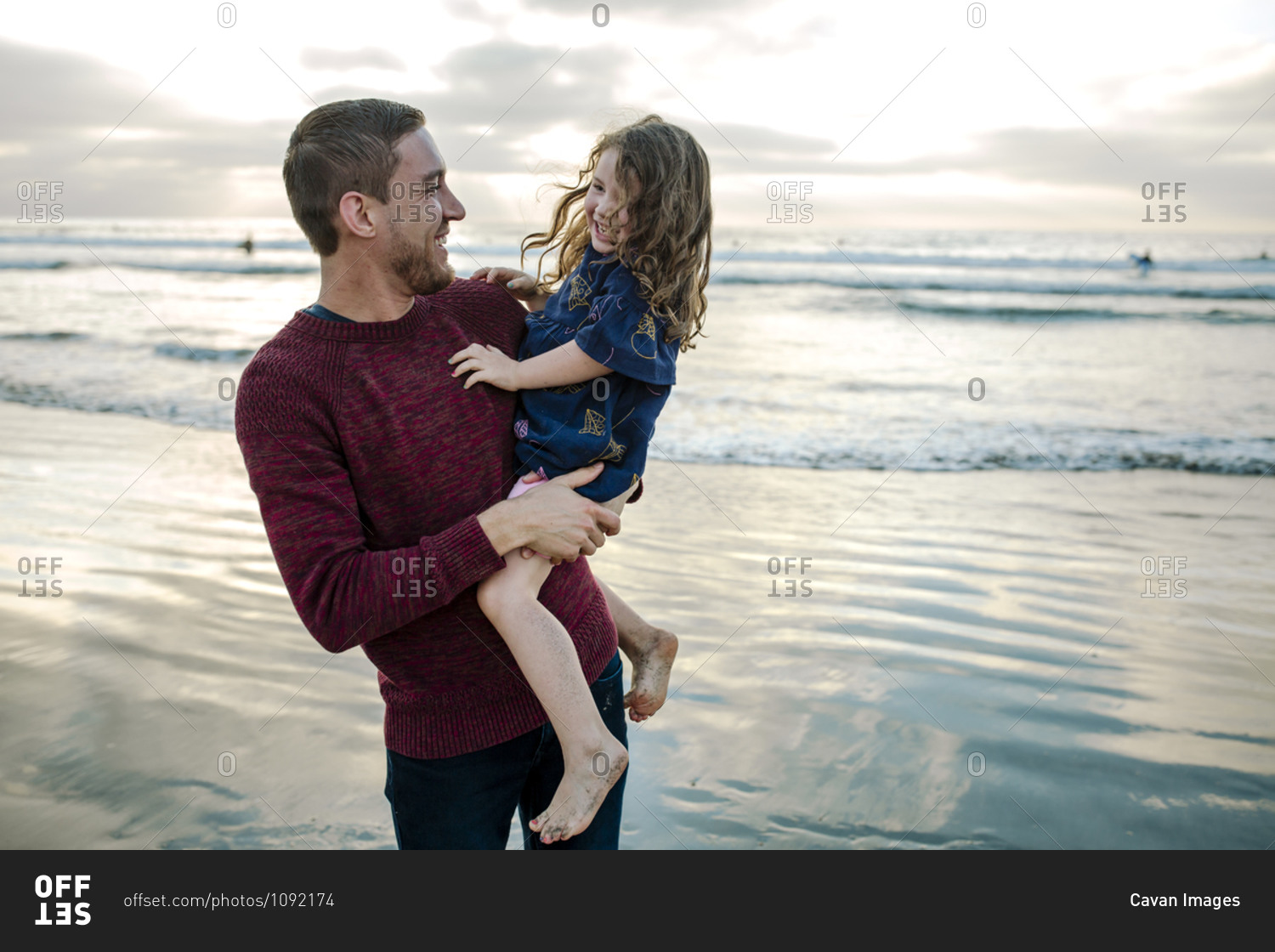 Young dad holding laughing bare-legged 3 yr old daughter at the ocean