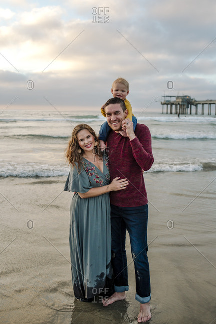 Joyful couple with baby on dad's shoulders on the beach near pier