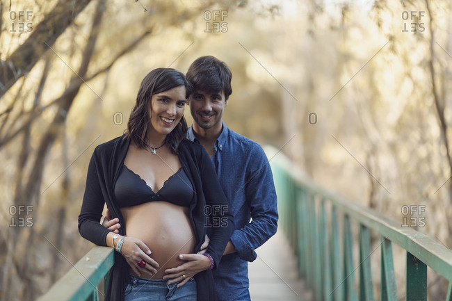 Portrait of a happy couple embracing woman's pregnant belly on a footbridge in autumn