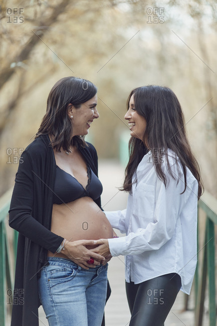 Two women laughing while embracing pregnant woman's belly on a footbridge in autumn