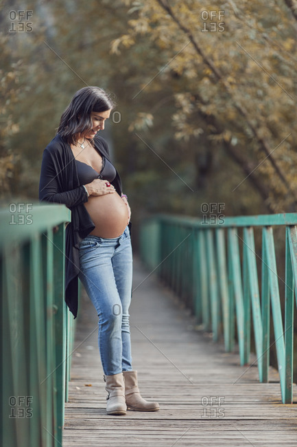 A pregnant woman standing on footbridge looking down at her belly in fall